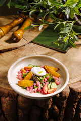 A vibrant bowl filled with assorted vegetables, poached egg, and potatoes rests on a rustic wooden table. Fresh greens and tomatoes add a pop of color to the dish.