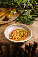 A pasta dish in a simple bowl sits on a wooden table, surrounded by greenery. The meal features rich sauce and hints of herbs, showcasing a cozy dining experience.
