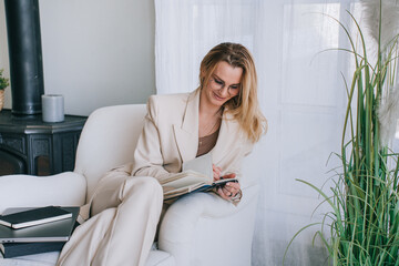 Seated comfortably by a large potted plant, a woman writes in her journal, immersed in thought and surrounded by quiet elegance.