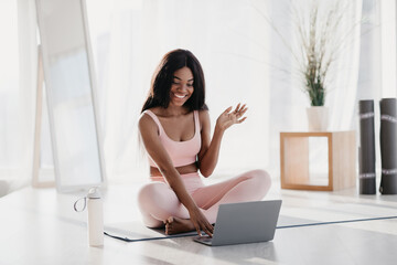 A young African American woman sits on a fitness mat in her living room, smiling as she greets her online personal trainer. She is dressed in sports clothes, preparing for a yoga session.
