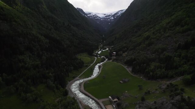Aerial view of a powerful river snaking through a valley of verdant green, flanked by dark forested mountains capped with snow, Buarbreen Glacier, Odda, Vestland, Norway.
