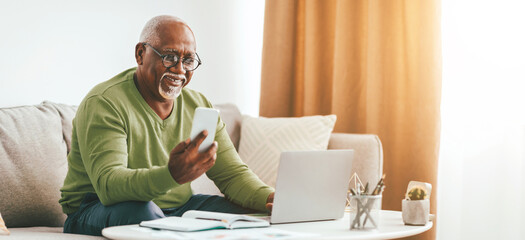 A senior man with glasses smiles as he holds a smartphone, engaged in a video call. He is seated on a sofa in a cozy living room setting with a laptop and plants nearby.
