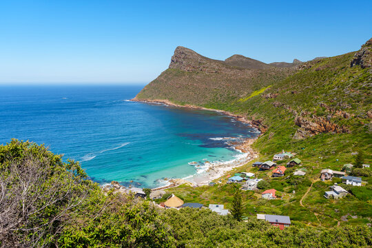 Breathtaking view of Smitswinkel Bay with turquoise Atlantic waters, sandy beach, quaint cottages in lush green hills, and Judas Peak in the background. Cape Peninsula, Cape Town, South Africa.