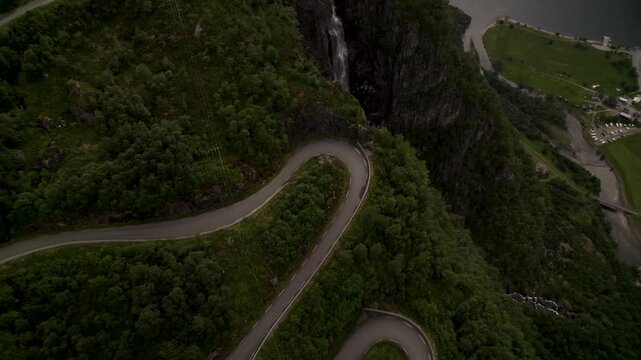 Aerial view of a winding road cutting through lush green mountains beside a fjord, creating a stark contrast with the dark water, Lysebotn, Rogaland, Norway.