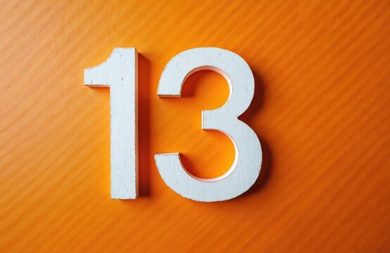 White wooden number 13 floats on bright orange background. Number is crafted from wood with distressed white paint finish. It sits against textured orange surface creating bold graphic element.