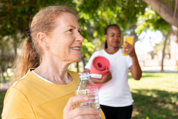 Senior woman hydrating after outdoor yoga class, embracing healthy active lifestyle in park with friend