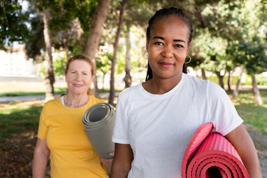 Diverse women friends carrying yoga mats for outdoor fitness session, enjoying an active healthy lifestyle in a park