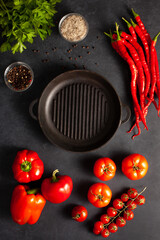 Top view of fresh tomatoes, red bell peppers, chili peppers, parsley, salt, peppercorns, and cast iron grill pan on dark stone background. Perfect for healthy cooking and recipe design.