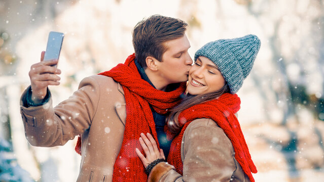 A young couple enjoys a winter day outdoors. They are bundled up in warm coats and scarves while sharing a loving kiss as they take a selfie. Soft snowflakes fall gently around them.