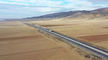 Aerial view of a long highway cutting through arid fields near mountains, highlighting the contrast between the grey road and brown landscape, Tuz, Ankara, Turkiye.