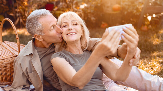 A couple of seniors share a loving moment as they relax on a picnic blanket in a park. They take a selfie together, surrounded by the warmth of a sunny day and nature.