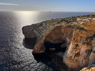 Dramatic view of the iconic Blue Grotto complex on the southern coast of Malta, captured against the sun's glare (backlighting). The high contrast emphasizes the deep shadows and the texture of the li