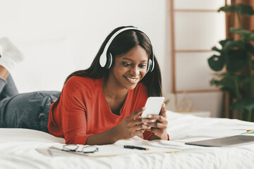 A young woman lies on her bed wearing headphones, smiling at her phone. She has notes and a laptop...