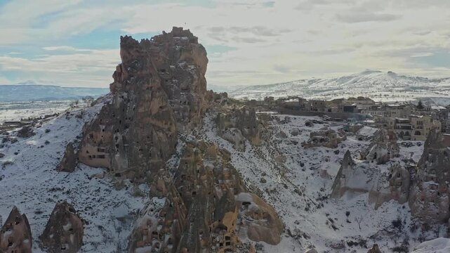 Aerial view of Uchisar Castle, a tall rock formation covered in snow, contrasting with the village houses below, Goreme, Cappadocia, Turkiye.