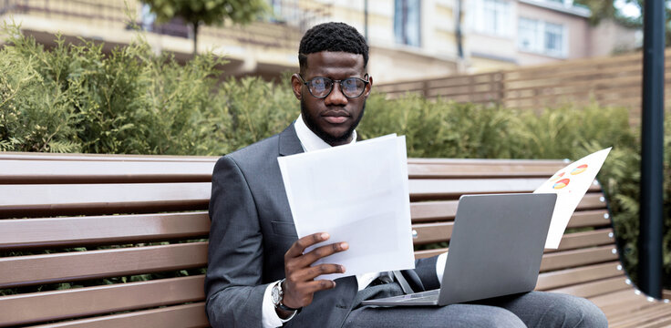A businessman in a suit sits on a park bench, analyzing papers while working on a laptop. The setting features greenery and urban buildings in the background, showing a serene working environment.