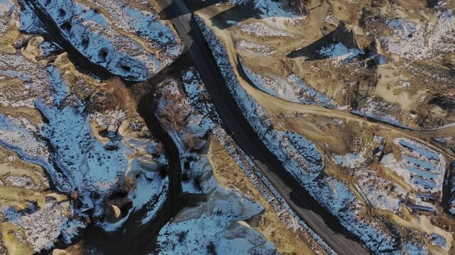 Aerial view of a winding road cutting through the snow dusted, rugged landscape of Cappadocia, the unique rock formations creating a stark contrast, Goreme, Cappadocia, Turkiye.