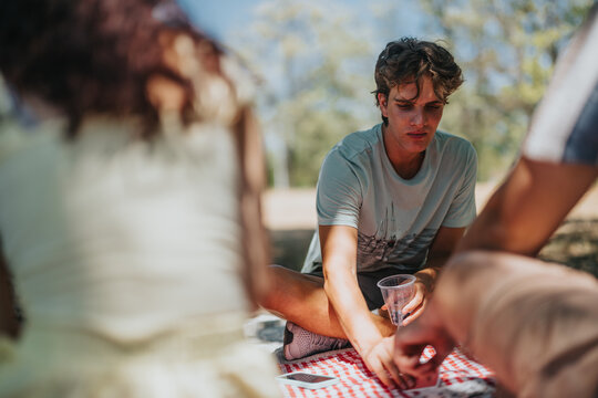 A group of friends sits on a red and white checkered blanket in a sunny park, sharing snacks and drinks. One man reaches to grab something as others relax and chat.