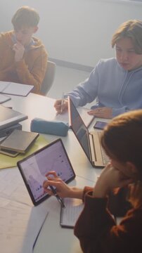 Concentrated Students Collaborate Around Shared Table, Combining Digital Tools and Handwritten Notes. Concept of Teamwork, Creative Problem Solving, and Blended Learning in Classroom. Vertical Shot.