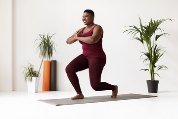 A motivated plus size African American lady is exercising in a fitness studio. She is doing lunges on a yoga mat, smiling and looking at the space around her while engaging in her workout routine.