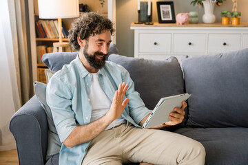 Smiling man waving during video call on digital tablet in living room