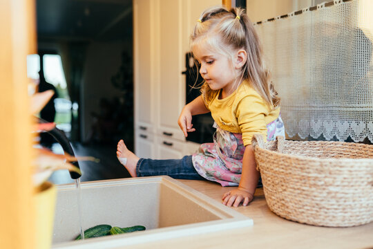 Toddler girl sitting on counter watching water running over cucumbers in kitchen sink