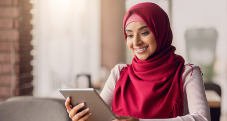 A woman wearing a red hijab smiles as she interacts with a tablet in a warm cafe. Soft light fills the space, creating a welcoming atmosphere for her engaging moment.