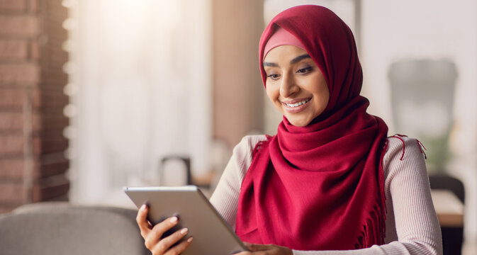 A woman wearing a red hijab smiles as she interacts with a tablet in a warm cafe. Soft light fills the space, creating a welcoming atmosphere for her engaging moment.