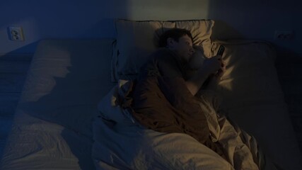 Man struggling with insomnia scrolling phone in bed at night, tired man unable to sleep, glowing smartphone screen reflecting on face