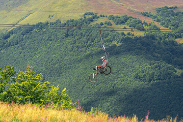 A mountain biker and their bicycle ascend a heavily forested mountain range via a chairlift on a bright, sunny day.