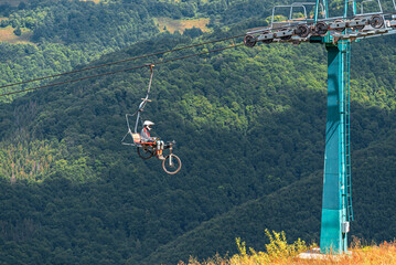 A mountain biker and their bicycle ascend a heavily forested mountain range via a chairlift on a bright, sunny day.