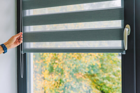 A woman opens black roller blinds on a window in a room. A woman's hand pulls a cord, closing the roller blinds in the room.
