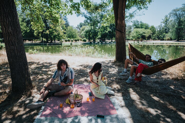A group of friends enjoys a warm afternoon in a park, sharing snacks on a checkered blanket beside a calm lake. One person sits, others relax in a hammock under shaded trees.