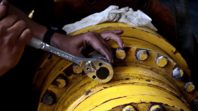 ​A mechanic's hands are actively using a ratchet wrench to tighten or loosen bolts on a worn, yellow industrial final drive housing during heavy equipment maintenance.