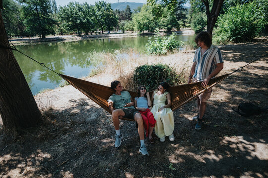 A group of friends lounges in a striped hammock between trees at a lakeside park, soaking up sunshine and conversation in a peaceful outdoor setting.