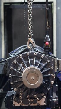 Automatic transmission running on test bench, lifted by a chain hoist during complex machinery assembly or maintenance in a workshop.