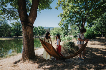 A group of friends in casual summer outfits hang a brown hammock between trees beside a tranquil lake in a park. The scene captures leisure, conversation, and carefree outdoor relaxation.