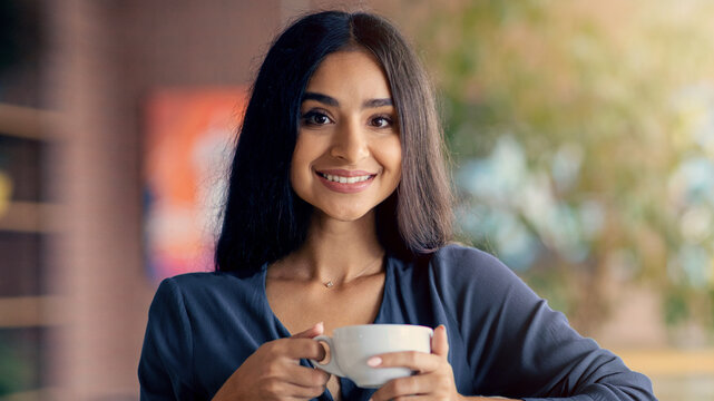 A young woman with dark hair smiles while holding a cup of coffee in her hands. She is in a warm, inviting cafe filled with natural light, creating a cheerful atmosphere in the morning.