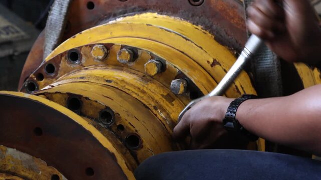 ​A mechanic's hands are actively using a ratchet wrench to tighten or loosen bolts on a worn, yellow industrial final drive housing during heavy equipment maintenance.