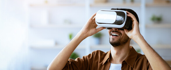 A young man joyfully explores virtual reality in a well-lit room filled with plants and modern furniture. His expression shows enthusiasm as he interacts with the VR headset.