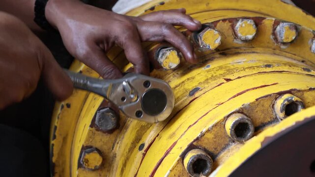 ​A mechanic's hands are actively using a ratchet wrench to tighten or loosen bolts on a worn, yellow industrial final drive housing during heavy equipment maintenance.