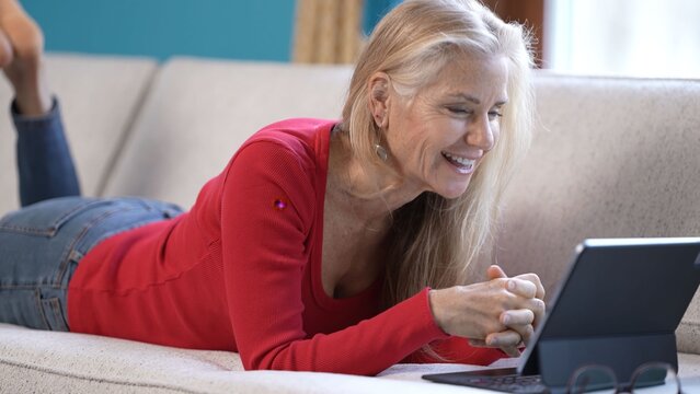 A middle-aged woman lies on her stomach on a couch, reading good news on her tablet computer. She shows expressions of surprise and happiness. - Powered by Adobe