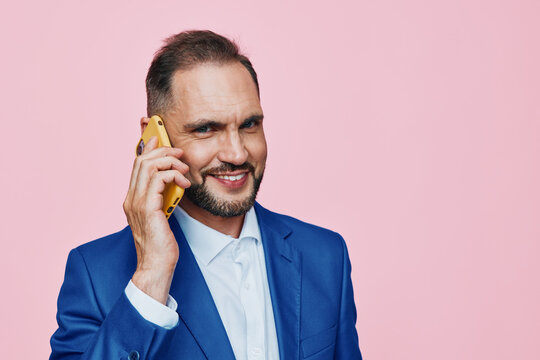 A confident business professional on a bright pink background, wearing a blue suit and white shirt, smiling while talking on a yellow phone, conveying an approachable mood.