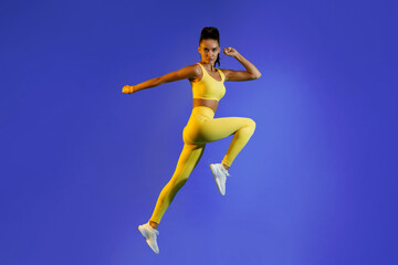 A woman leaps in mid-air, showcasing athleticism and grace. She wears a yellow sports outfit and white sneakers, set against a vibrant blue backdrop. The scene captures a sense of motion and fitness.