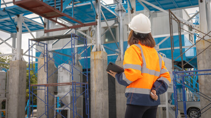 A construction site manager stands with a clipboard, wearing safety gear and observing ongoing work. The scene captures the collaboration and activity at a building site in clear daylight