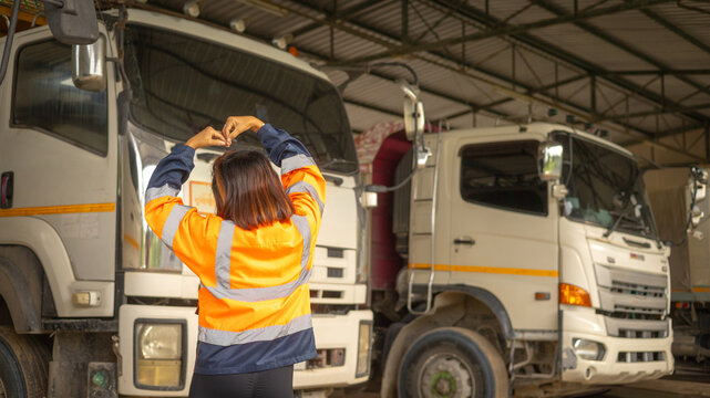 A worker in an orange safety jacket forms a heart shape with her hands in front of large trucks parked in a garage. The scene captures a moment of happiness in a busy workplace