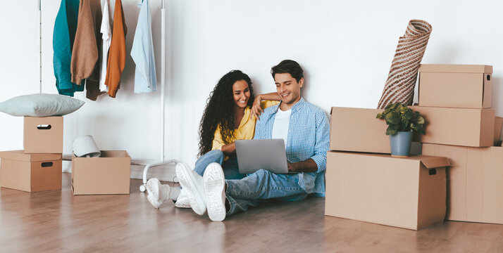 A young couple sits on the floor in a new home, smiling and looking at a laptop. Surrounding them are unpacked boxes and clothing hung on a rack. The atmosphere feels cozy and inviting. - Powered by Adobe