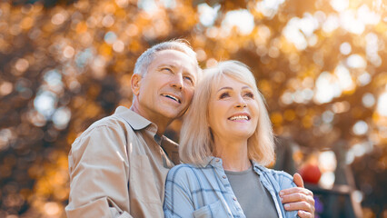 Fototapeta premium A joyful couple stands close together in a park, surrounded by colorful autumn leaves. They share a warm smile as sunlight filters through the trees, creating a serene atmosphere.