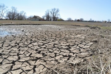 A dry, cracked landscape shows the effects of drought, with sparse vegetation and a distant house under a clear blue sky.