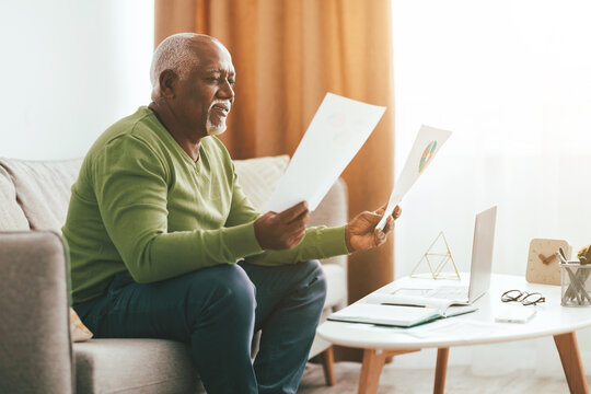 An older gentleman sits comfortably on a sofa, examining several papers in his hands. A laptop rests on a coffee table nearby, with sunlight streaming through the window. - Powered by Adobe
