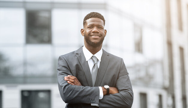 A man in a formal suit stands confidently with his arms crossed in front of a modern office building. He exudes professionalism and self-assurance in a bright, sunlit environment. - Powered by Adobe